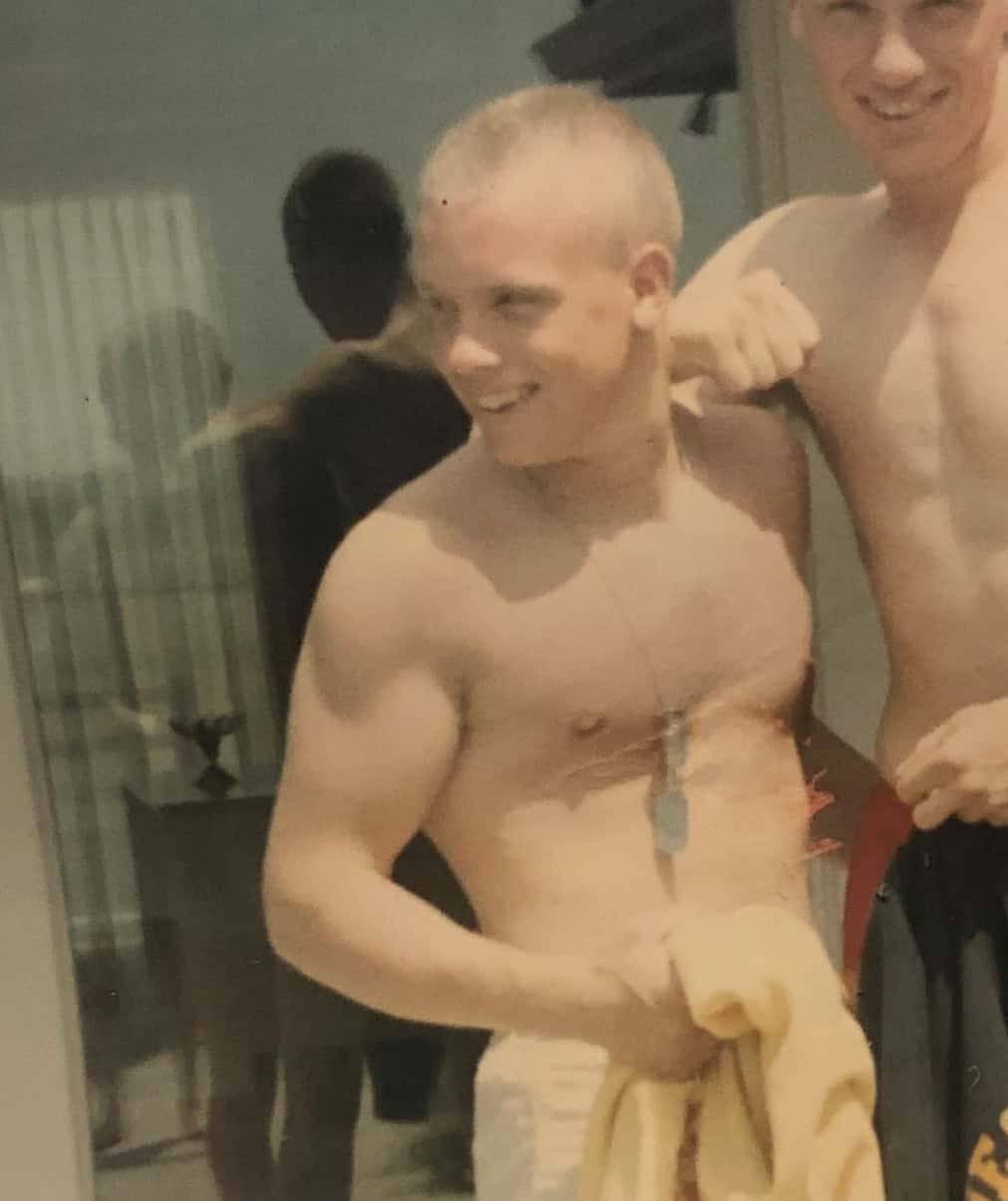 Young man with a shaved head smiling, shirtless, in a casual indoor setting.