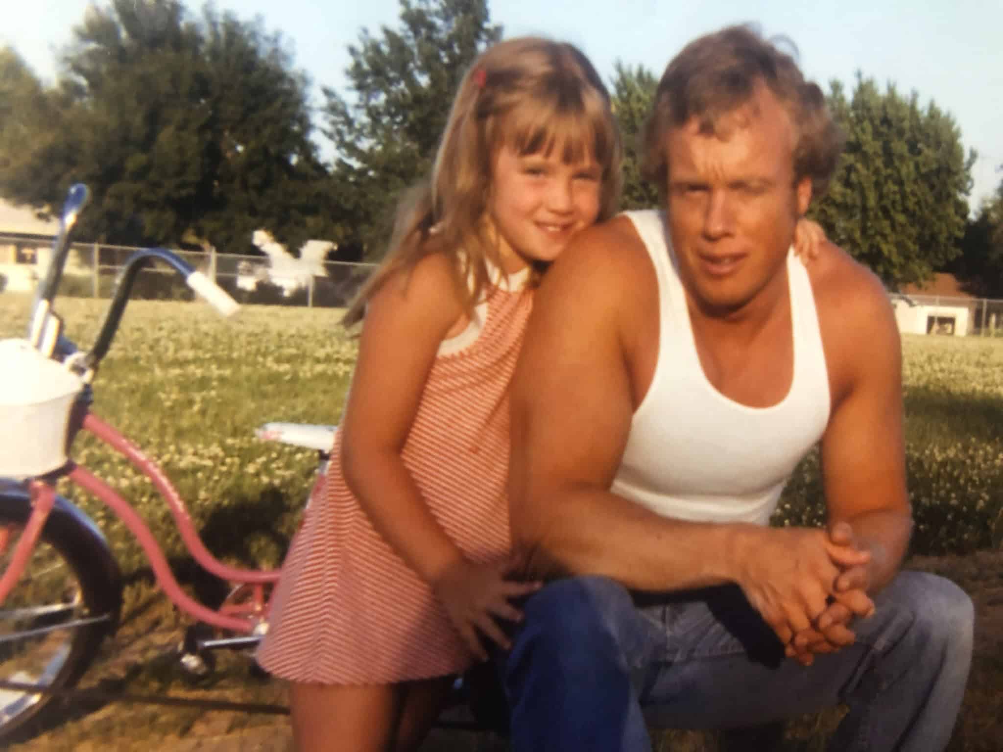 A young girl and a man sitting outdoors with a pink bicycle nearby, smiling.