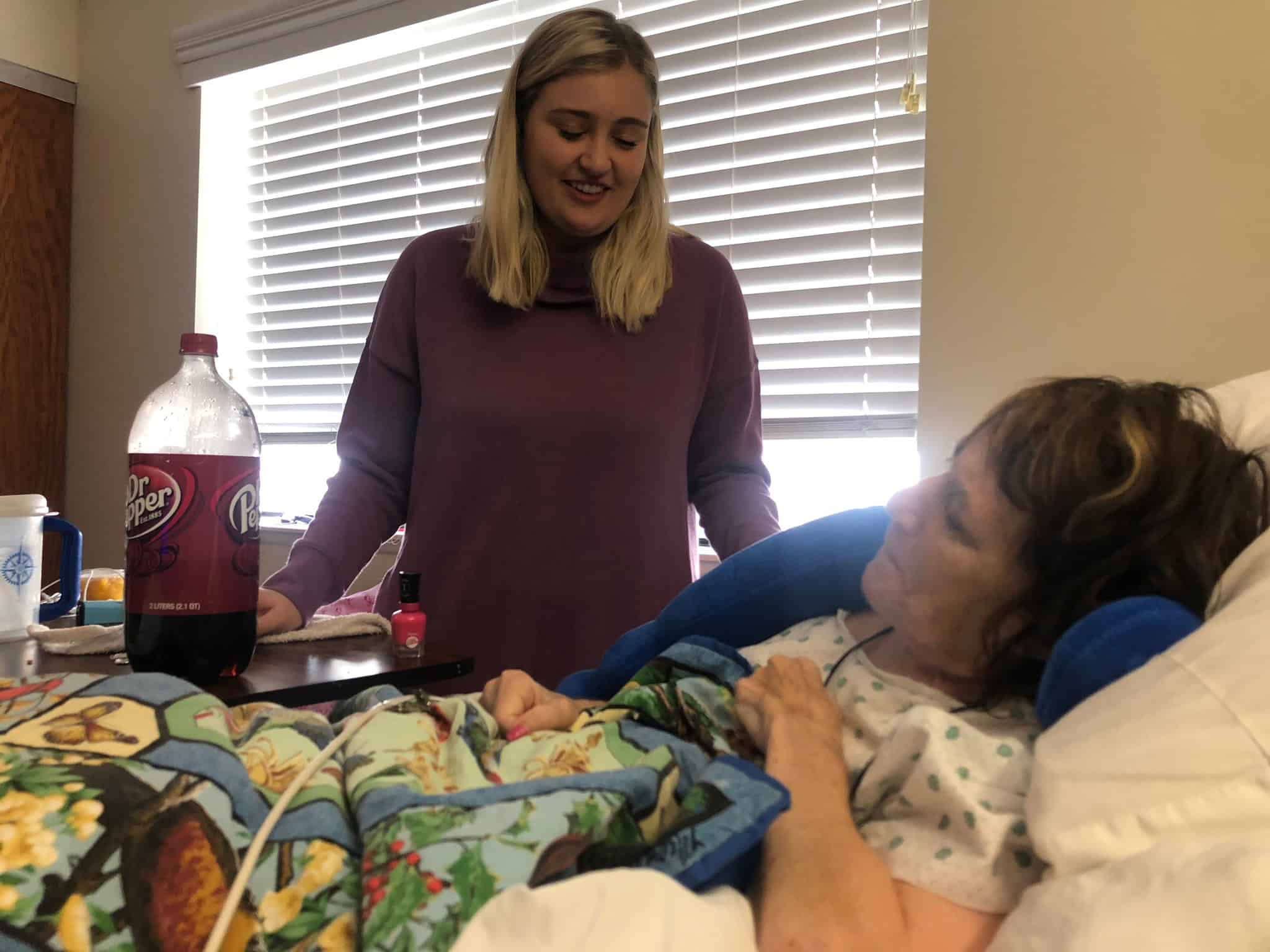 Young woman smiling at a patient in a hospital bed with colorful blanket and drinks nearby.